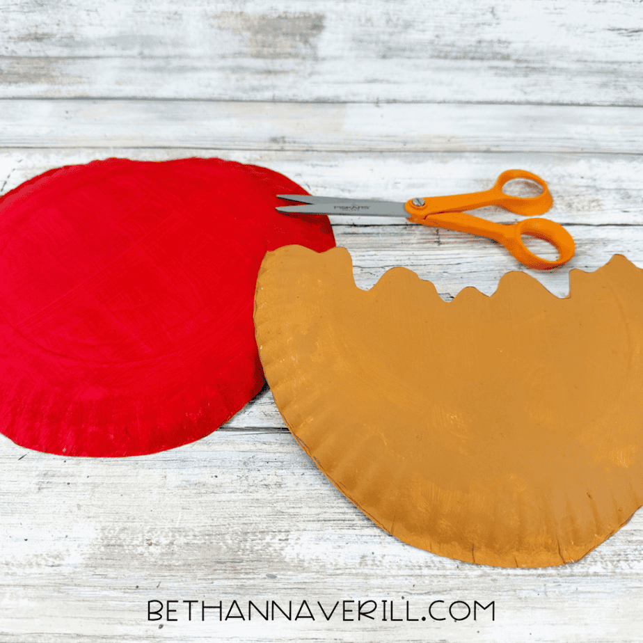 Red painted paper plate beside a caramel brown paper plate piece cut with a wavy edge and orange scissors.