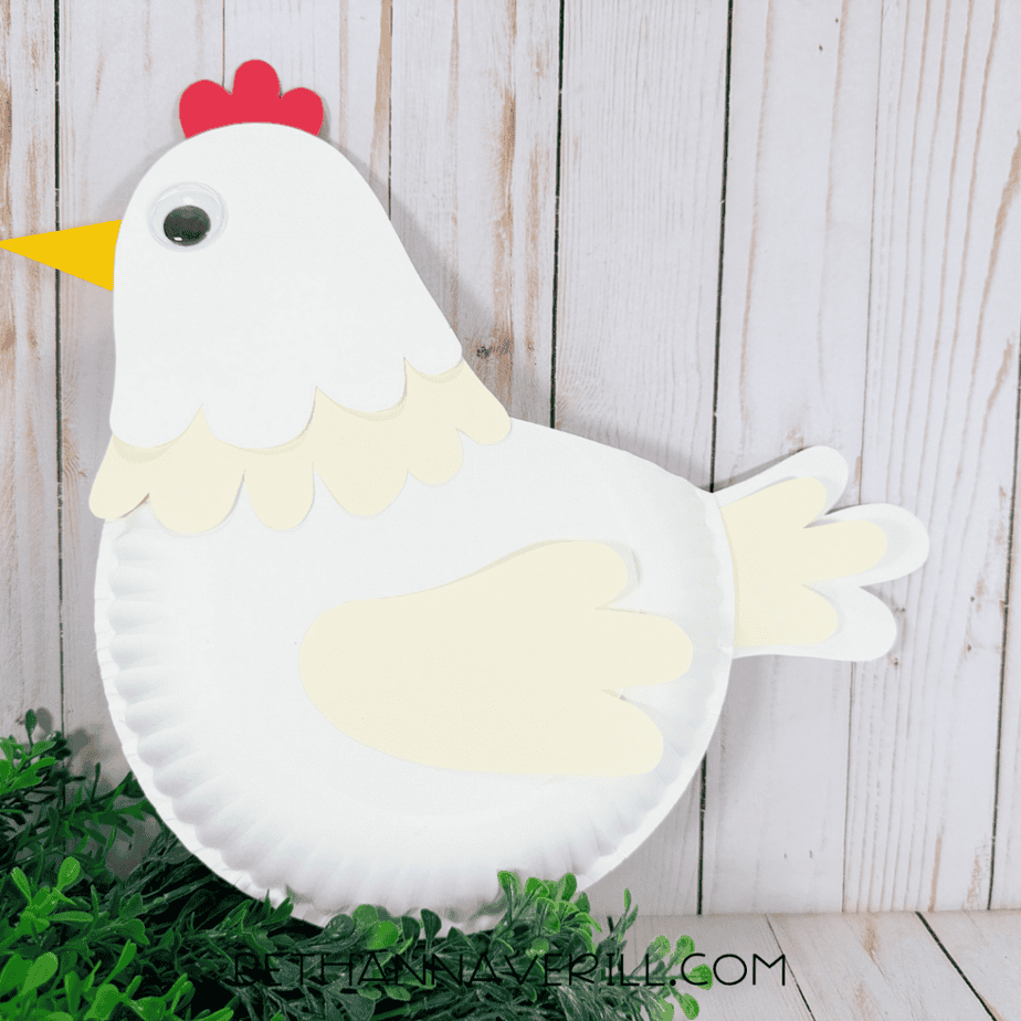 Finished paper plate chicken with a white body, white head, red comb, yellow beak, googly eye, cream wing, layered neck feathers, and tail feathers.