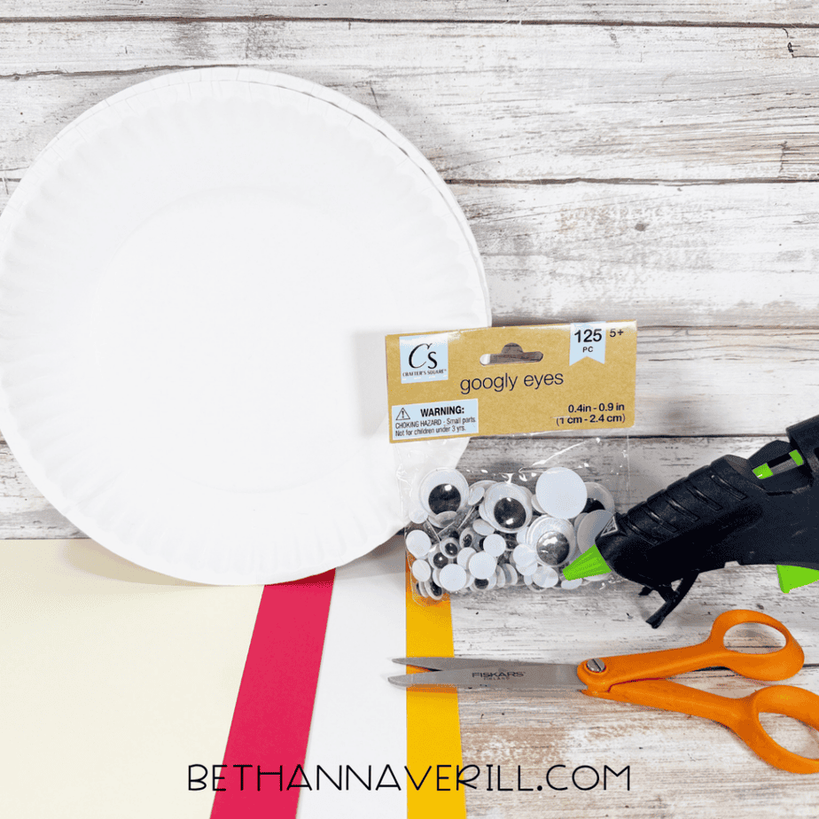 White paper plate with cream, red, yellow, and white paper, googly eyes, hot glue gun, and orange scissors arranged on a white wood surface.