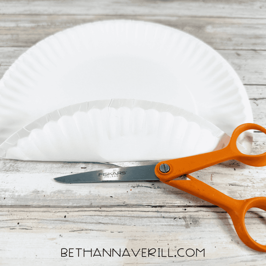 White paper plate being cut with orange scissors, with a curved strip removed from the top edge.