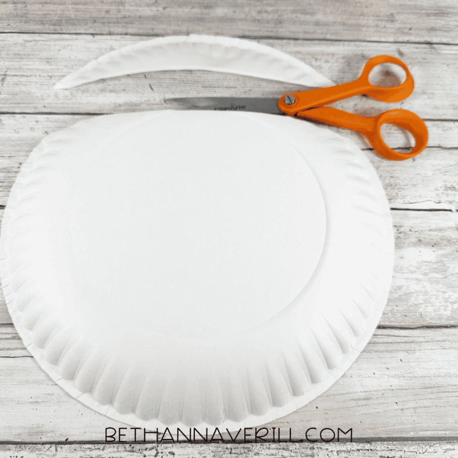 White paper plate with a curved strip cut away from one side, with orange scissors above it.