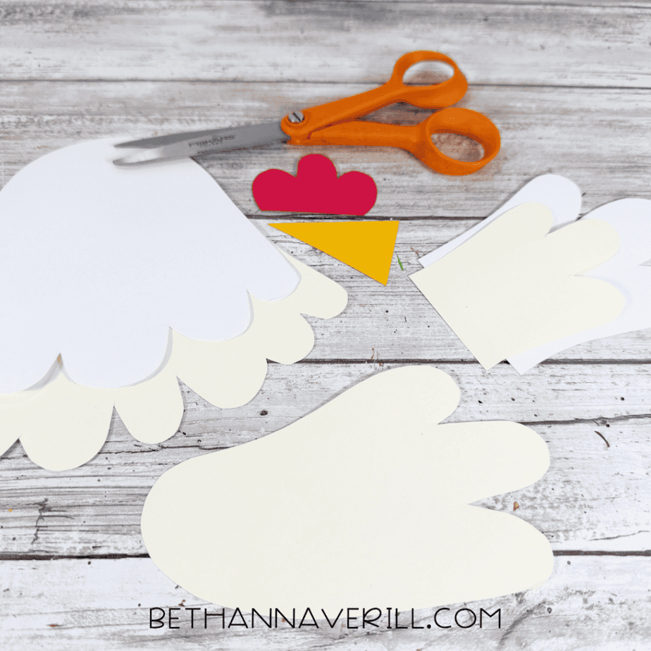 Cut paper pieces arranged on a white wood surface, including cream wing and feather shapes, white pieces, a red comb, and a yellow triangular beak, with orange scissors above.