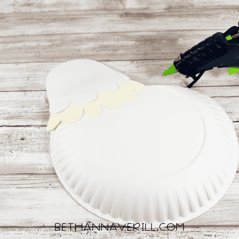 White paper plate with layered white and cream feather pieces attached near the top, with a hot glue gun nearby.