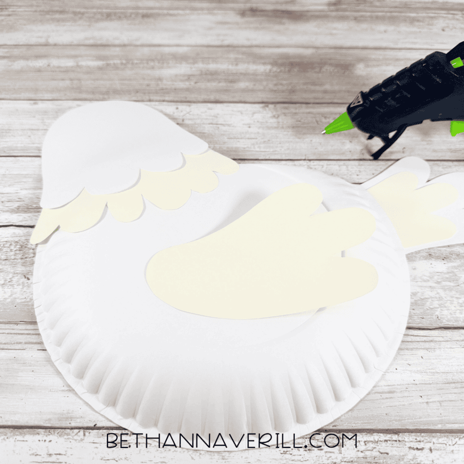 White paper plate with layered feather pieces at the top and cream wing and tail pieces positioned on the plate, with a hot glue gun nearby.
