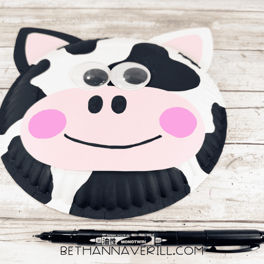 Paper plate cow face with black spots, googly eyes, pink ears and muzzle, pink cheeks, black nostrils, and a curved black smile, with a black marker below.