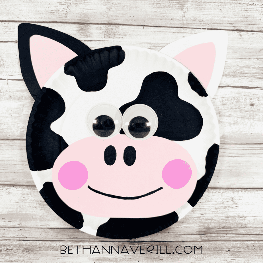 Finished paper plate cow face with black-and-white spots, pink-centered ears, large googly eyes, pink muzzle and cheeks, black nostrils, and a smiling mouth.