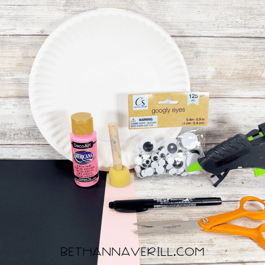 White paper plate with pink acrylic paint, foam dauber, googly eyes, hot glue gun, black marker, orange scissors, and sheets of black, pink, and white paper.