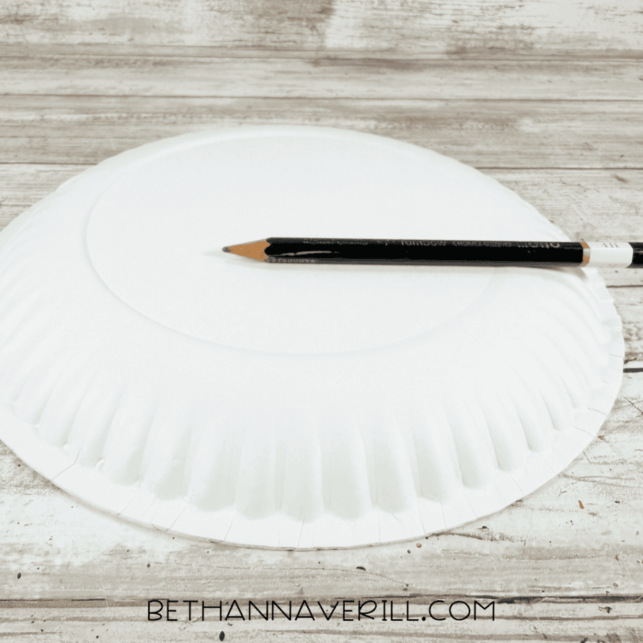 Upside-down white paper plate on a white wood surface with a black pencil resting across the center.