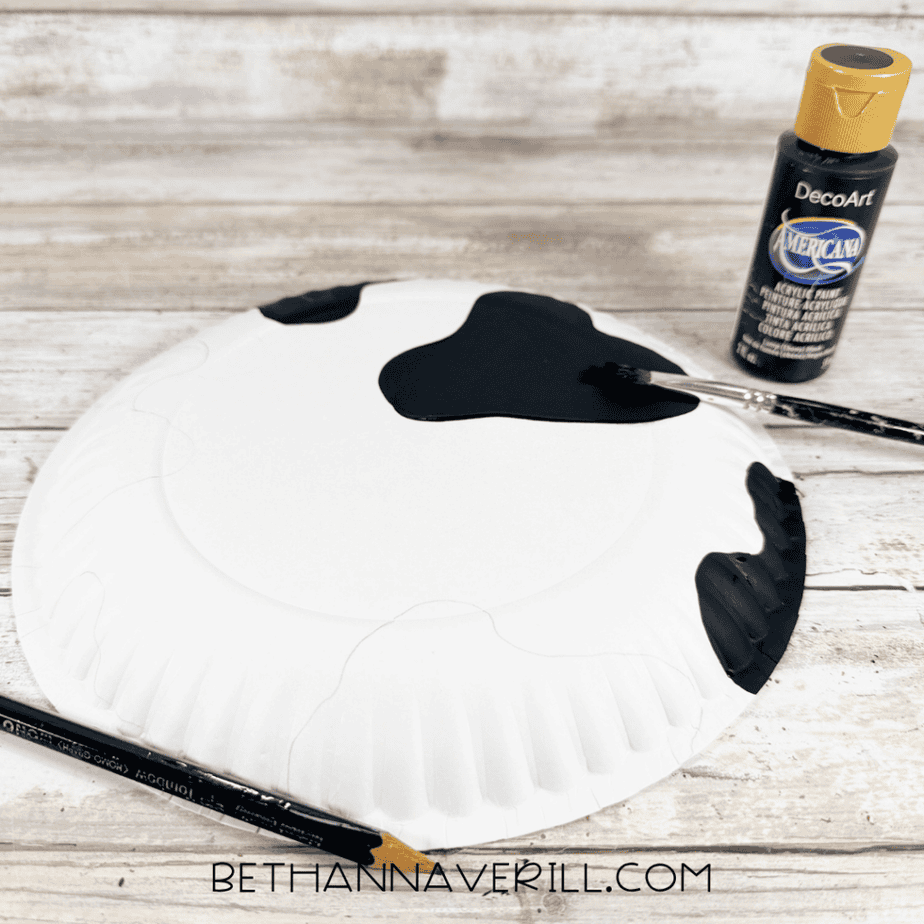 Black acrylic paint being brushed onto irregular spots on a white paper plate, with a black paint bottle beside it.
