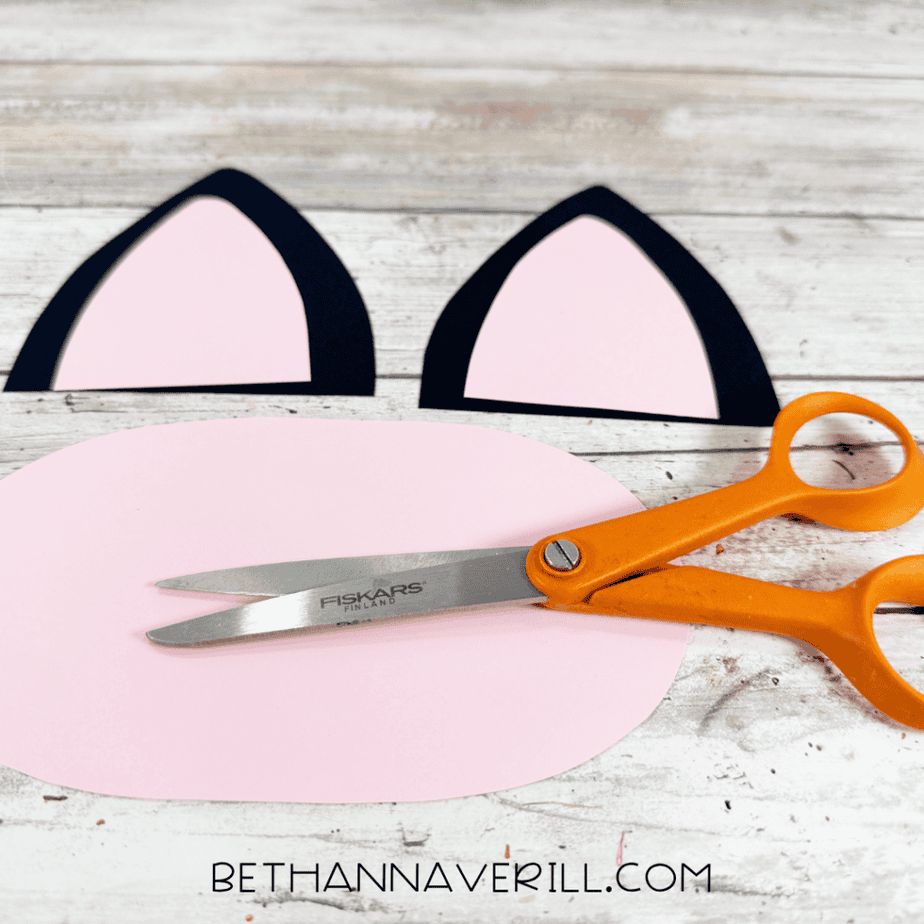 Two black paper ear shapes with pink centers and a large rounded pink paper shape arranged beside orange scissors.