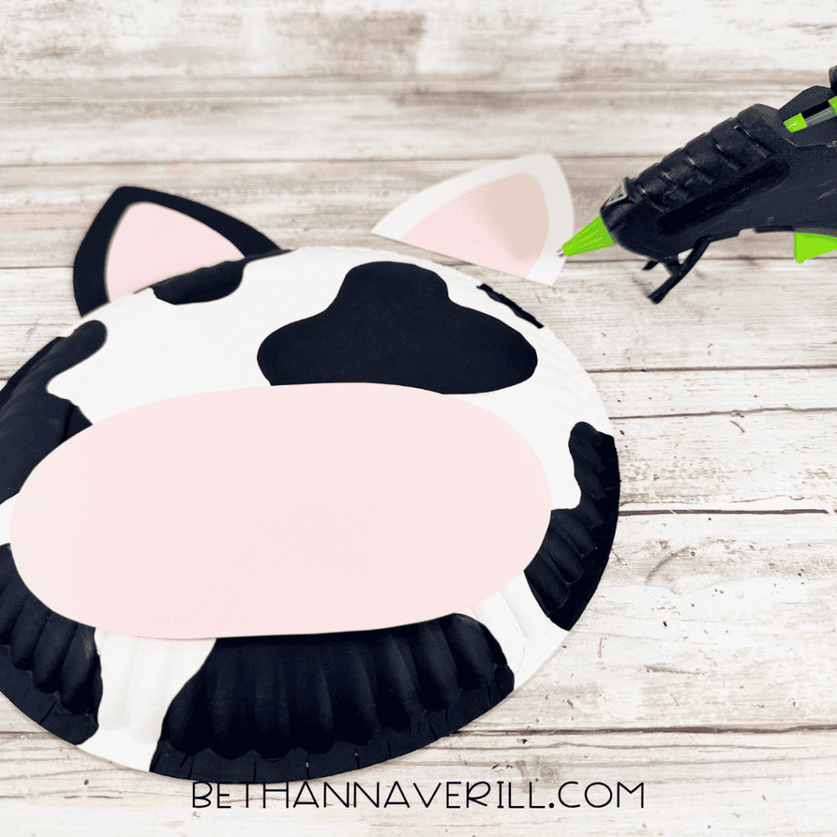 Black-and-white spotted paper plate with a large pink oval paper piece across the front and two pink-centered ear pieces above, with a hot glue gun nearby.