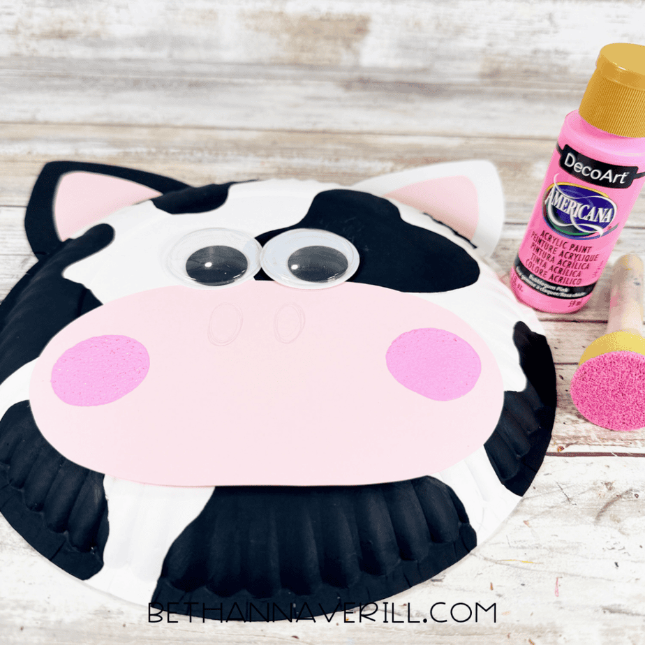 Paper plate cow face with black spots, pink-centered ears, large googly eyes, a pink muzzle, and two pink circles, beside pink paint and a foam dauber.