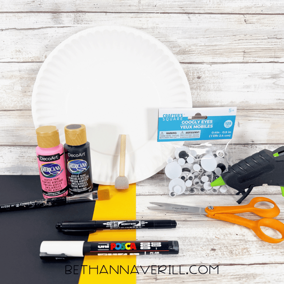 Craft supplies arranged on a white wood surface, including paper plates, black and pink paint, googly eyes, black and yellow paper, scissors, markers, brushes, and a hot glue gun.