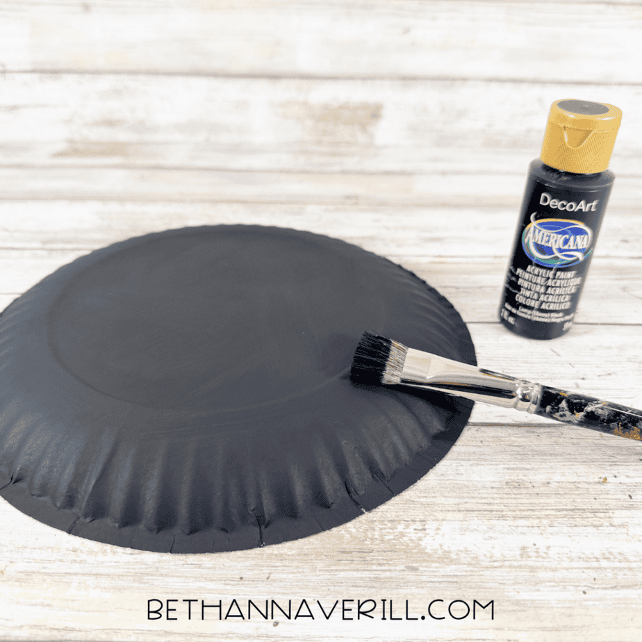 Paper plate painted black with a paintbrush and bottle of black acrylic paint beside it.