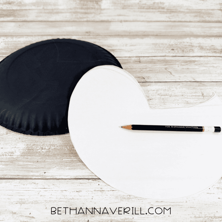Black painted paper plate beside a large white paper template with a curved bird body shape and a pencil.