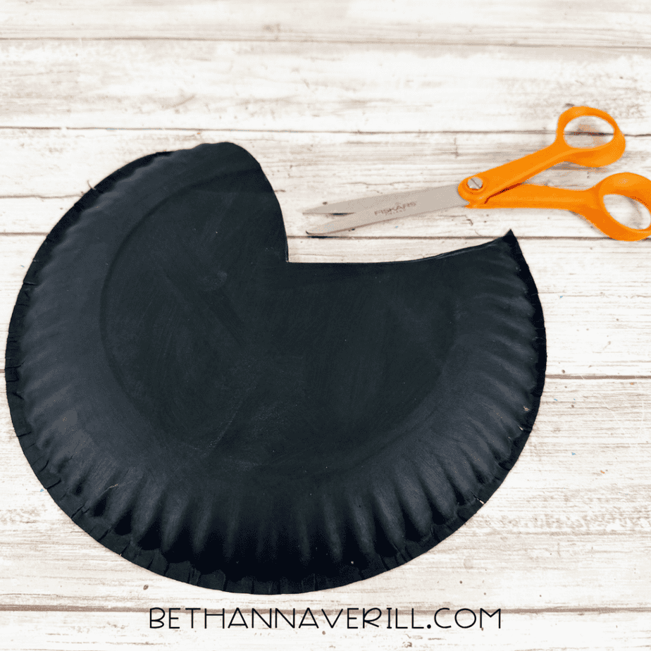 Black painted paper plate cut into a rounded bird body shape with an extended tail section, with orange scissors above it.