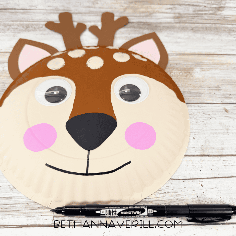 Paper plate deer face with antlers, ears, googly eyes, pink cheeks, cream forehead spots, black nose, and a finished curved mouth.