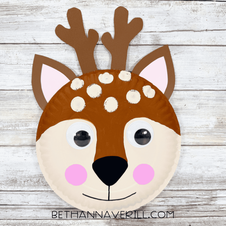 Completed paper plate deer craft with brown antlers, layered ears, googly eyes, cream spots, pink cheeks, and a black nose on a white wood background.