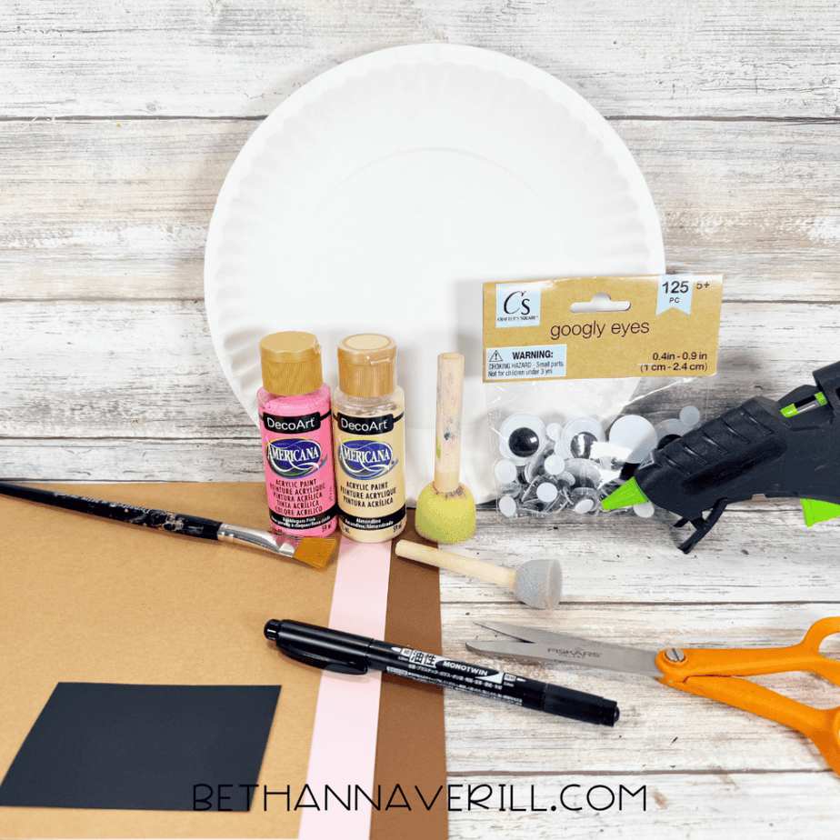White paper plate with brown, pink, and black cardstock, googly eyes, paint, brushes, scissors, marker, and hot glue gun arranged on a white wood surface.