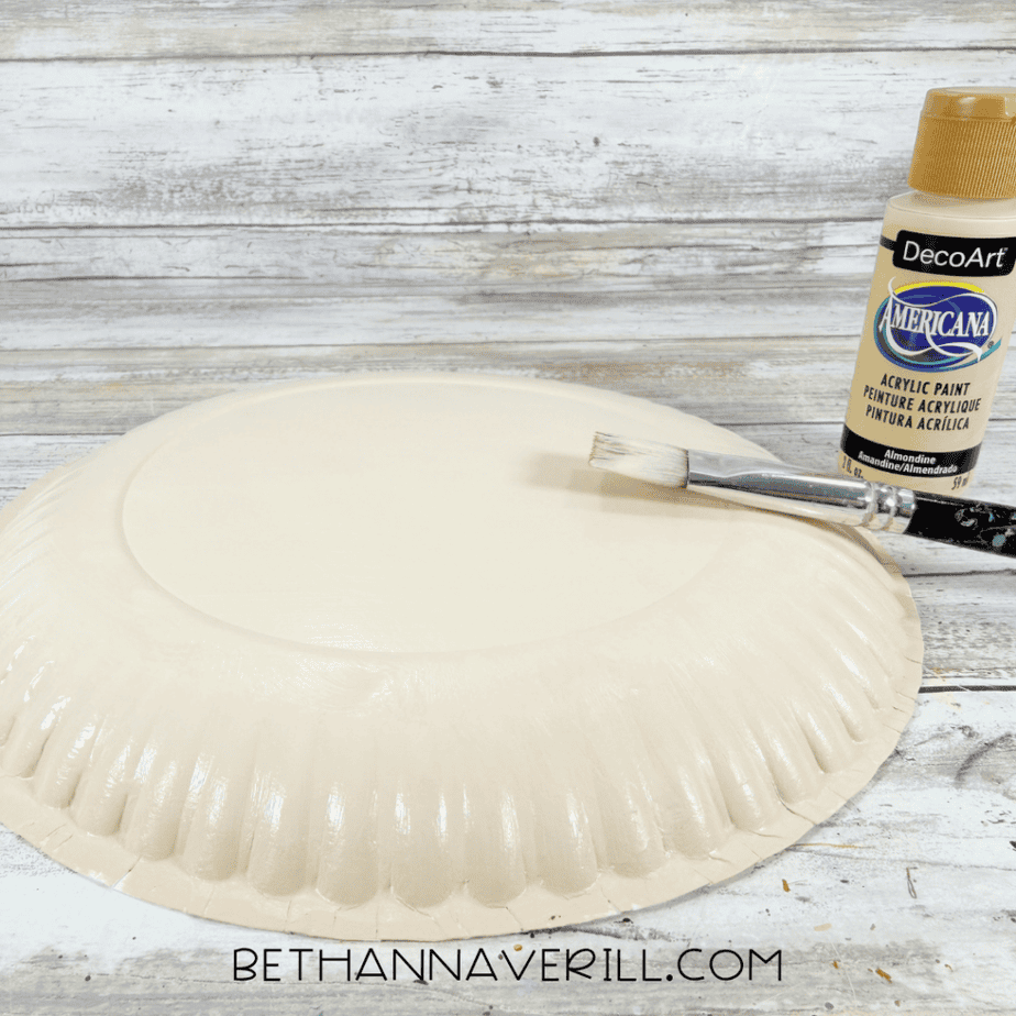 Paper plate painted a light cream color beside a bottle of acrylic paint and a paintbrush.