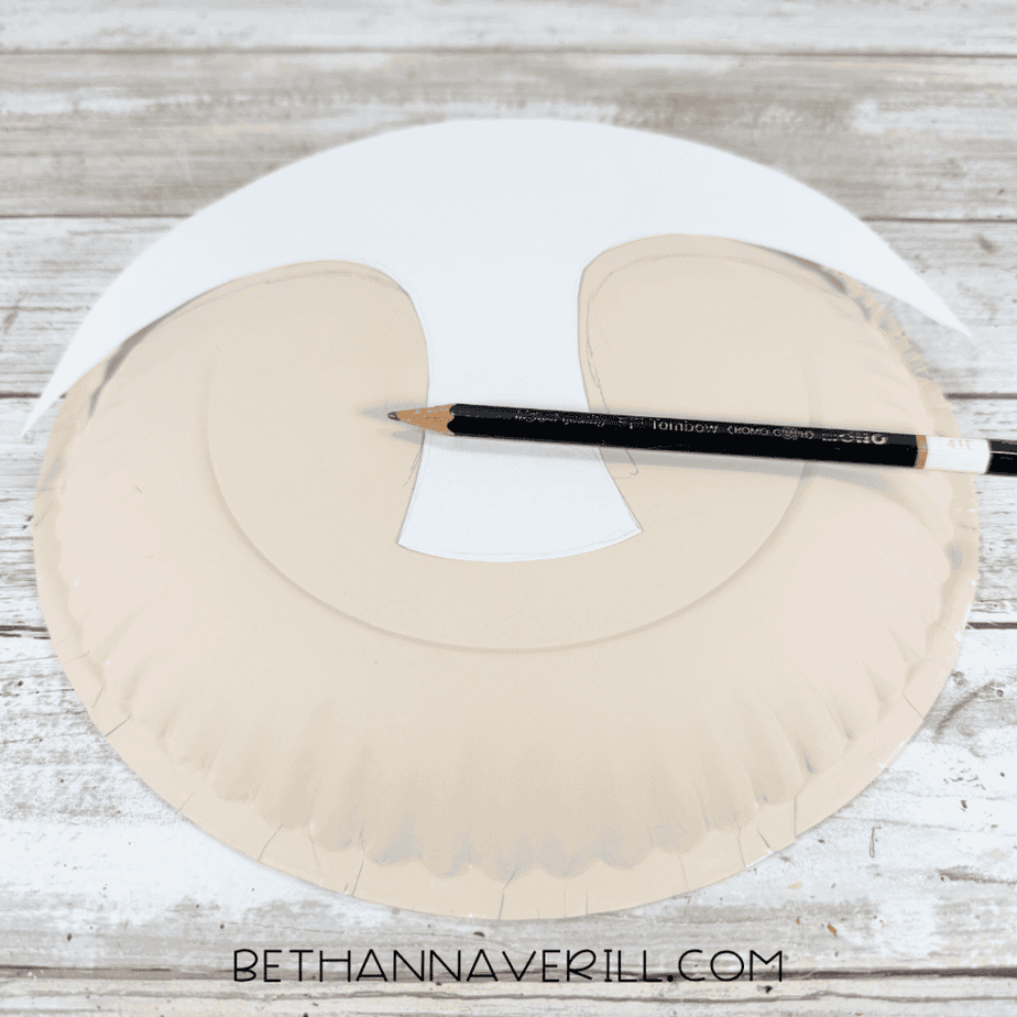 Cream paper plate with a paper template positioned over the top and center while a pencil marks the shape.