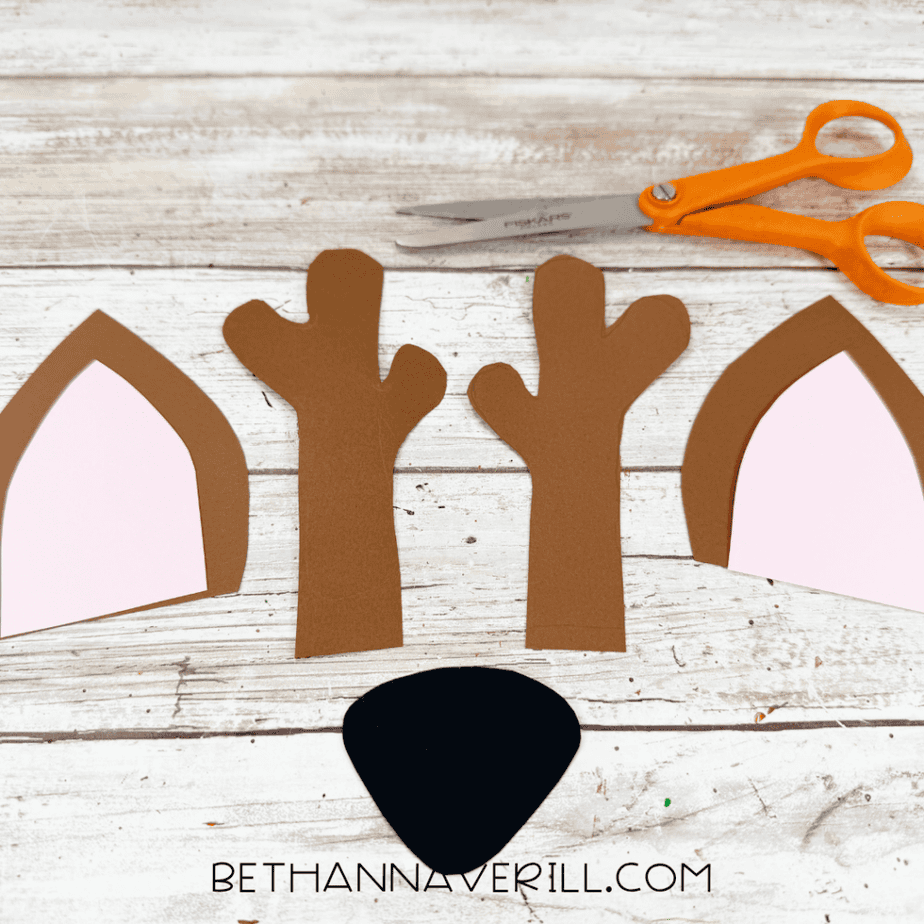 Cut brown antlers, layered brown and pink ears, and a black nose arranged beside orange-handled scissors.