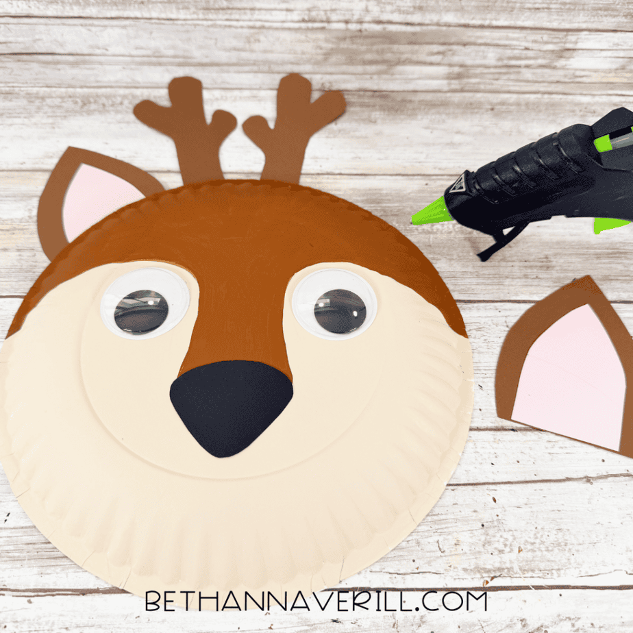Partially assembled paper plate deer face with antlers, one ear, googly eyes, cream face sections, and a black nose beside a hot glue gun.