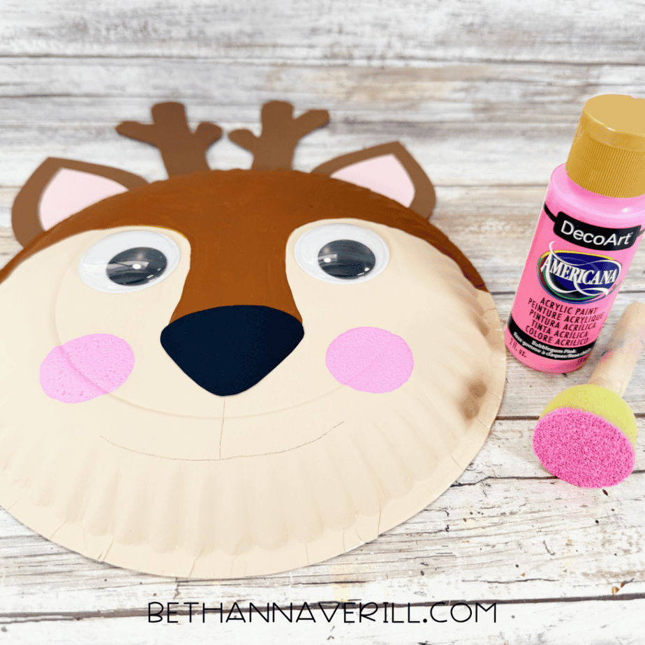 Paper plate deer face with antlers, both ears, googly eyes, black nose, pink cheeks, and a lightly drawn mouth beside pink paint and a sponge brush.