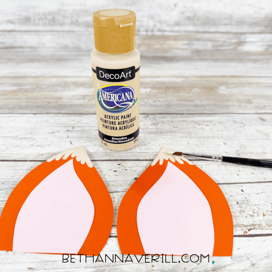Cream acrylic paint being added to the pointed tips of two orange and pink paper fox ears.