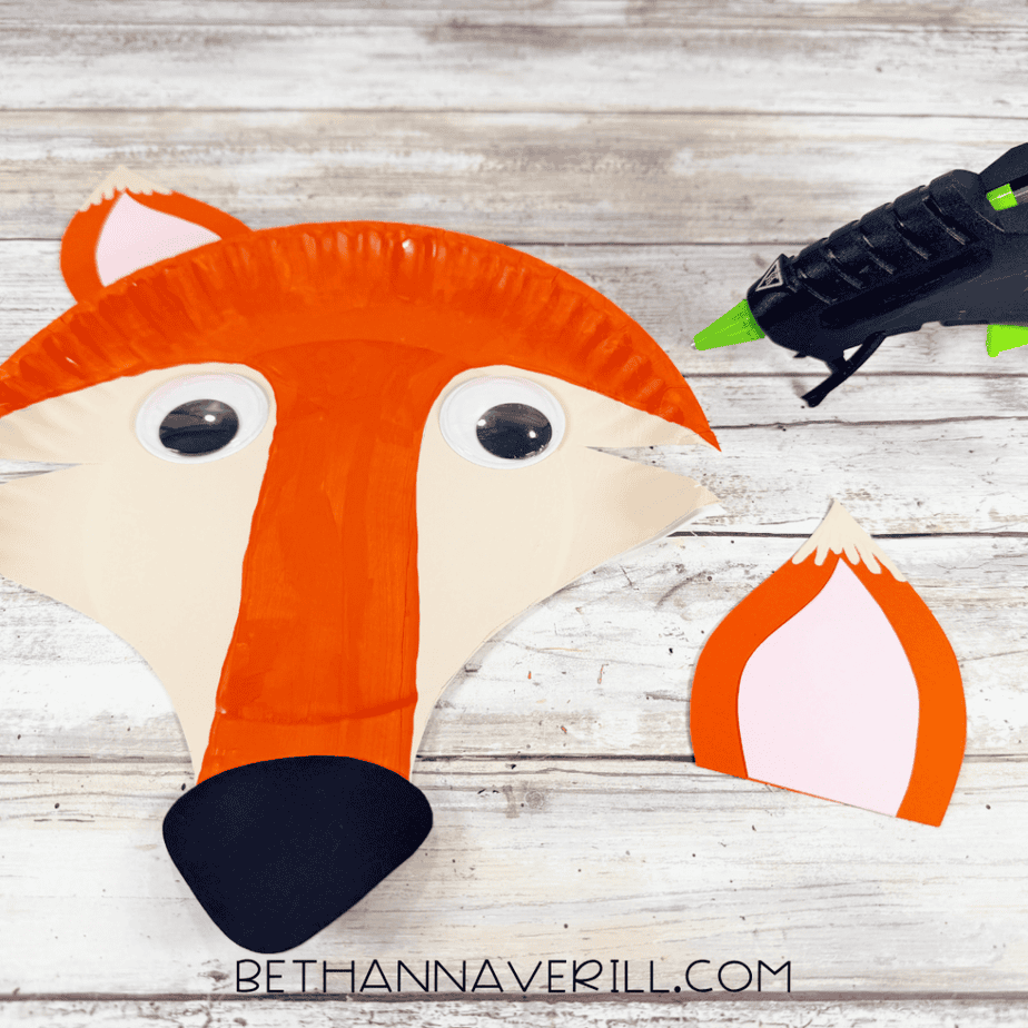 Partially assembled paper plate fox face with googly eyes, orange and cream painted sections, black nose and one paper ear attached.
