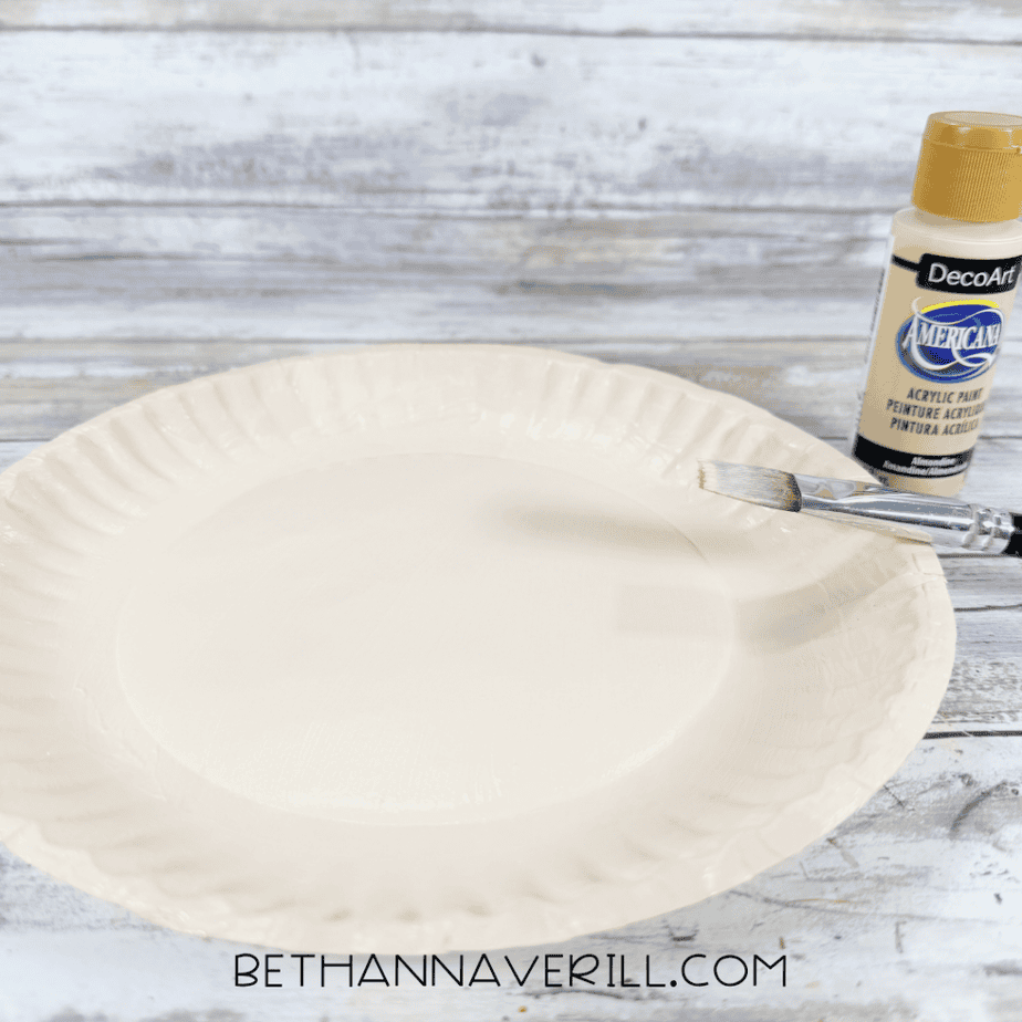 Paper plate being painted a light cream color with acrylic paint and a flat paintbrush.