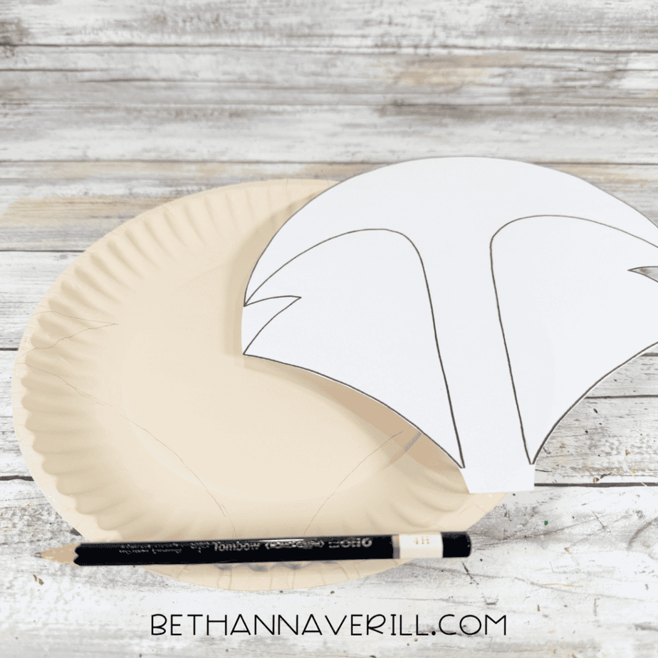 Paper fox face template positioned over a cream-painted paper plate with pencil outlines visible on the plate.
