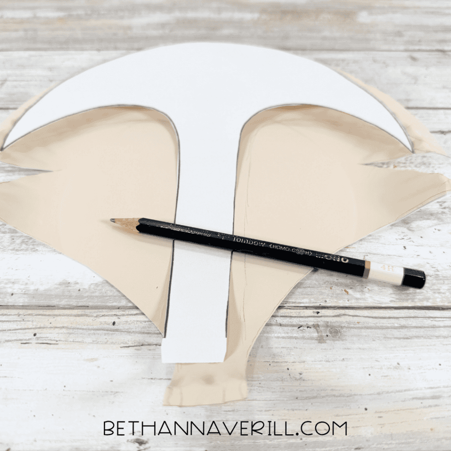 Paper fox face template laid over the cut paper plate with a pencil for tracing the center and upper face sections.
