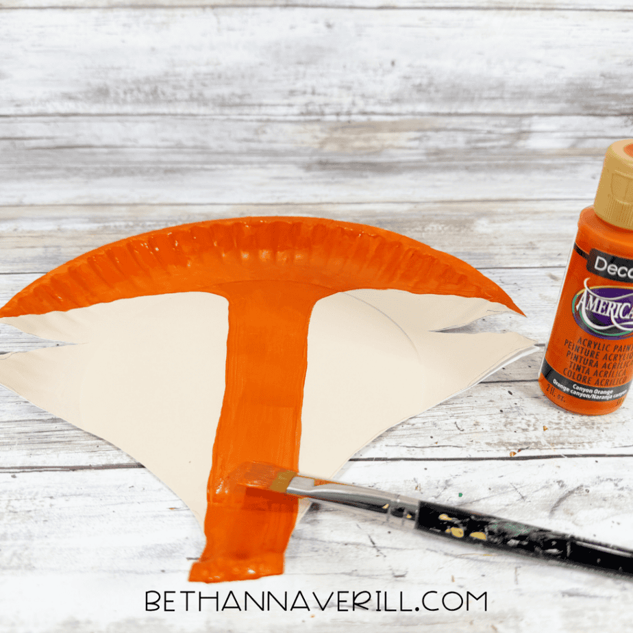 Orange paint being brushed onto the top and center of a cream paper plate cut into a fox face shape.