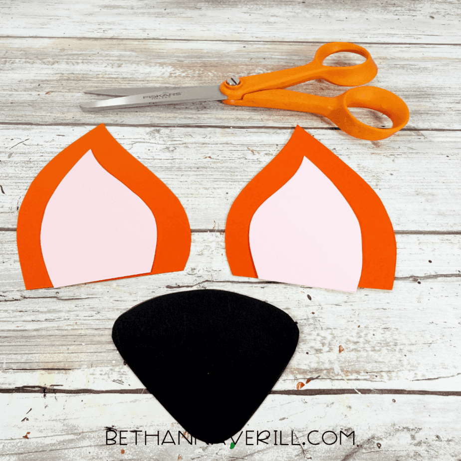 Two orange and pink paper fox ears and a black paper nose arranged beside orange scissors.