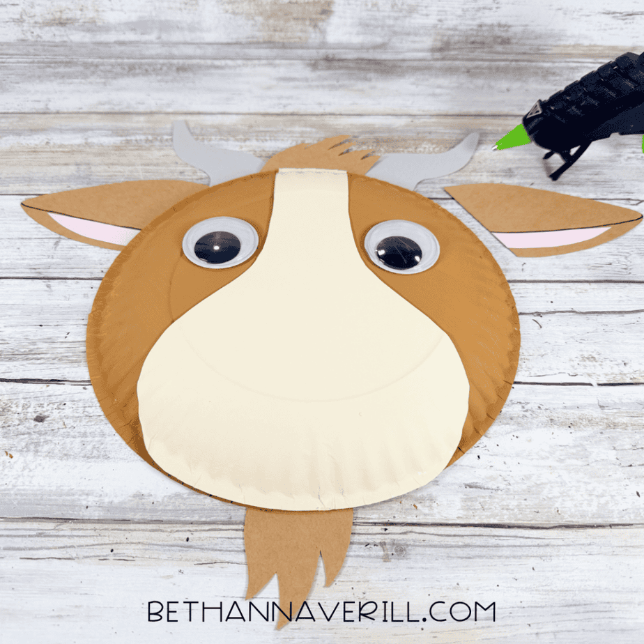 Partially assembled paper plate animal face with a brown head, cream muzzle, large googly eyes, brown ears, gray horns, brown hair, and a brown beard shape.