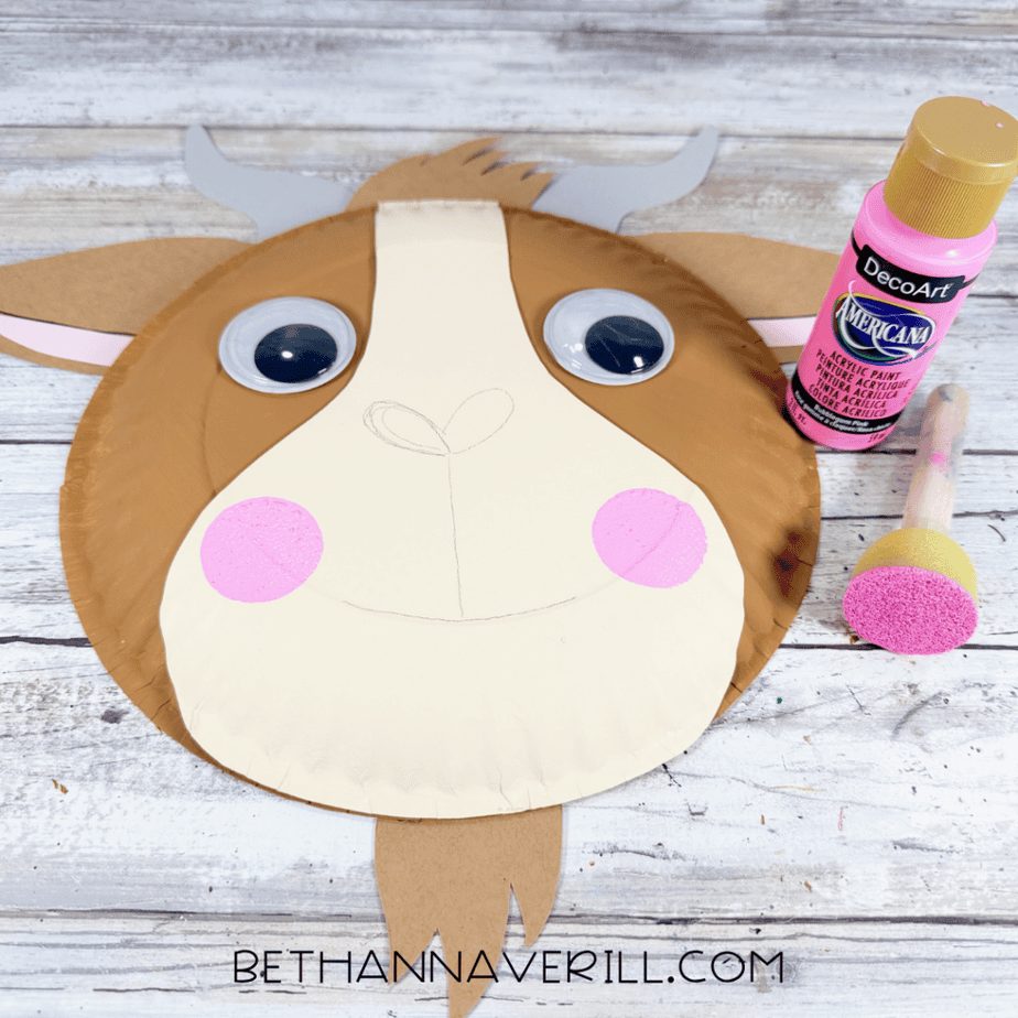 Paper plate animal face with pink circular cheeks added to the cream muzzle, beside a pink paint bottle and round foam dauber.