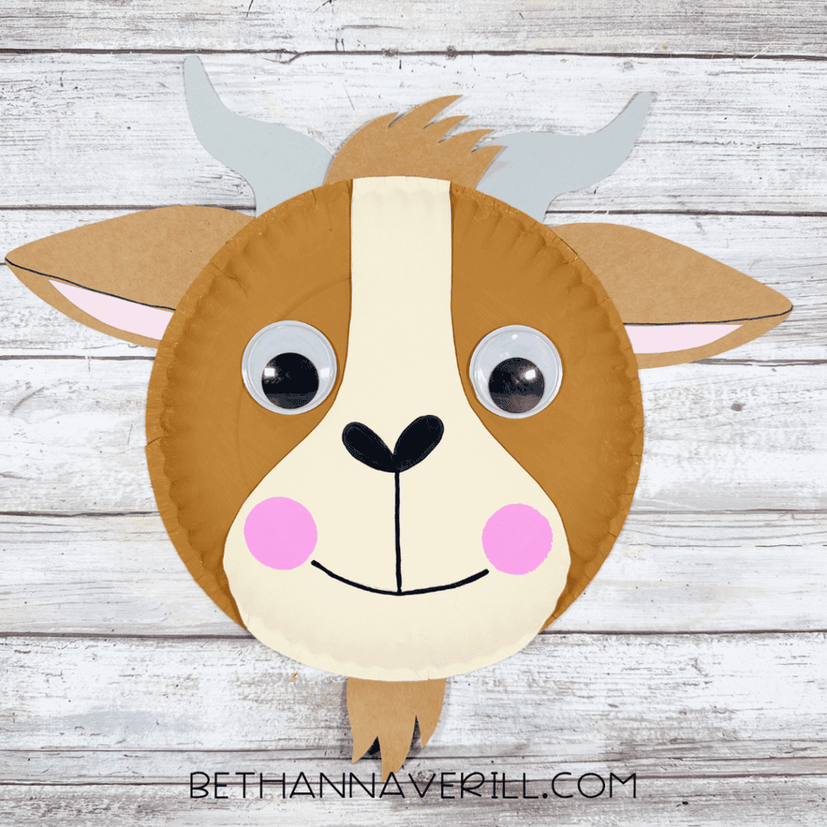 Finished paper plate goat face with brown and cream coloring, googly eyes, gray horns, brown ears with pink centers, pink cheeks, black nose and smile, and brown hair and beard pieces.