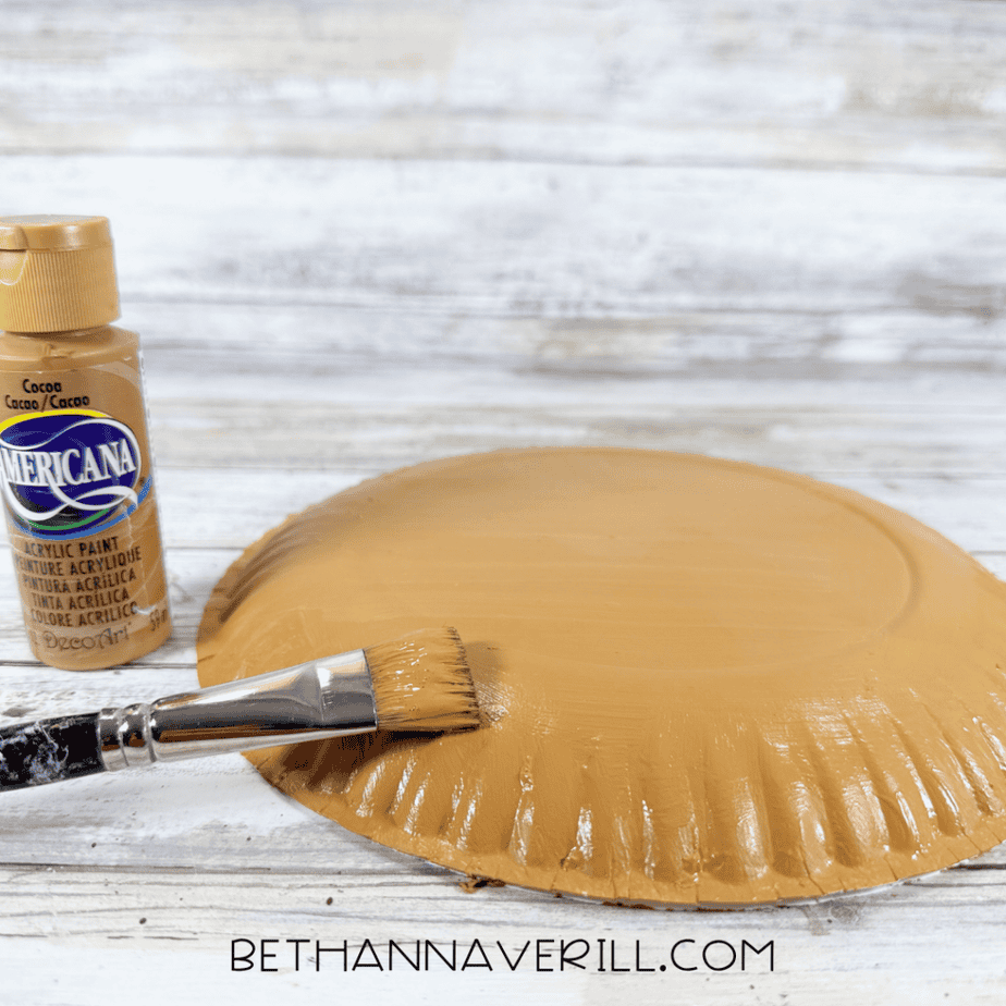 Paintbrush applying brown acrylic paint to the back of a paper plate on a white wood surface.