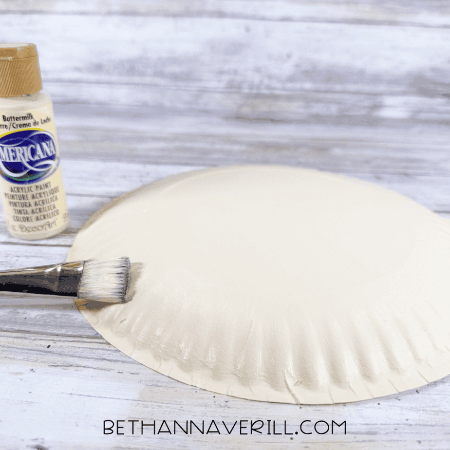 Paintbrush applying cream-colored acrylic paint to the back of a paper plate, with a paint bottle beside it.