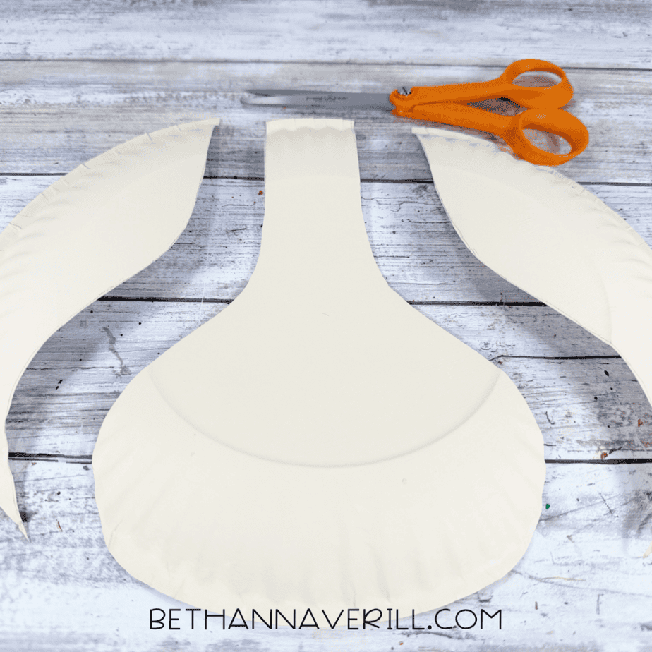 Cream-painted paper plate cut into a wide center shape with two curved side pieces removed, with orange-handled scissors above.