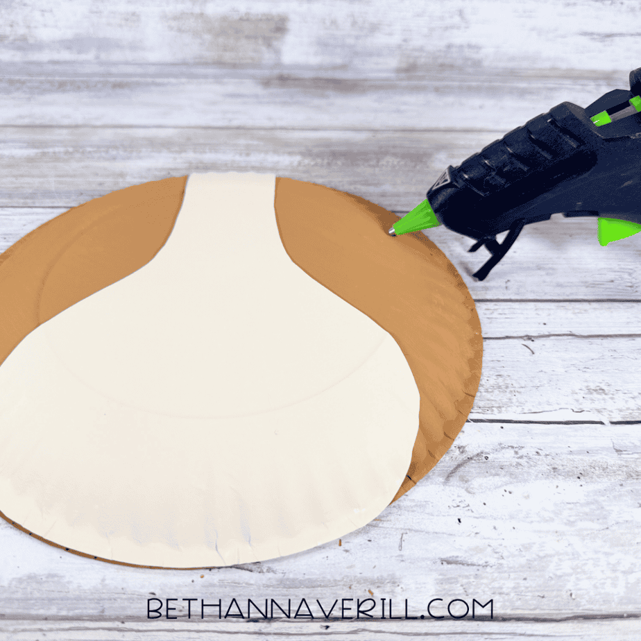 Cream paper plate piece positioned over a brown painted paper plate, with a hot glue gun beside the layered pieces.