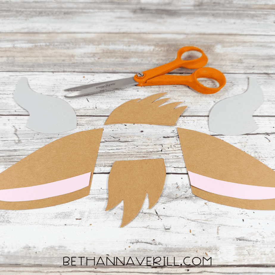 Cut paper pieces arranged on a white wood surface, including gray horn shapes, brown ears with pink centers, and brown hair and beard shapes, with scissors above.