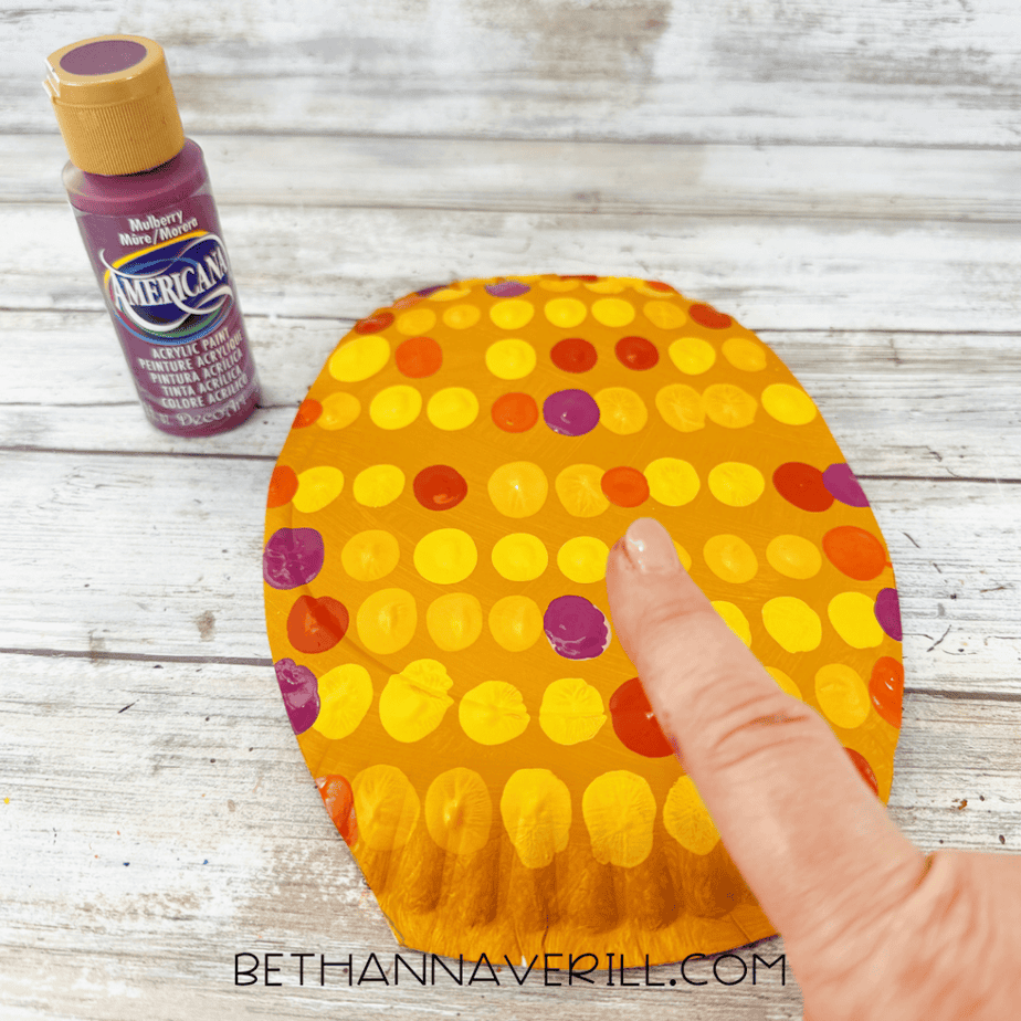 Golden yellow paper plate with yellow, orange, and purple paint dots as a finger adds purple paint, with a purple acrylic paint bottle beside it.