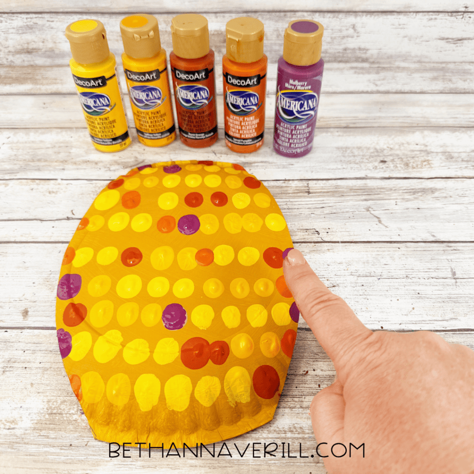 Golden yellow paper plate covered with yellow, orange, and purple paint dots with five acrylic paint bottles arranged above it.