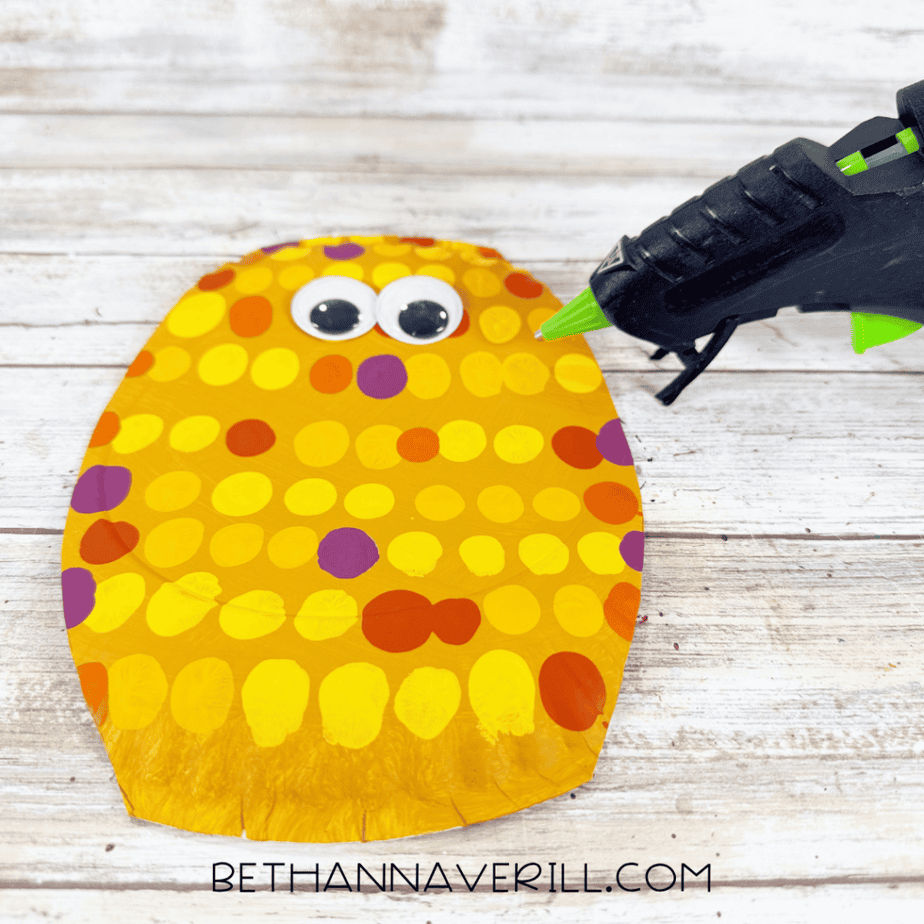 Dotted golden yellow paper plate with two googly eyes attached near the top and a hot glue gun beside it.