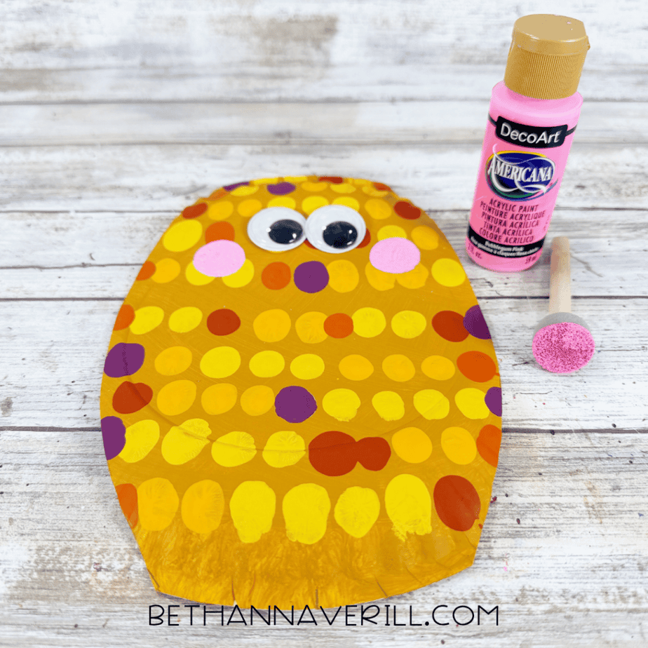 Dotted paper plate with googly eyes, pink painted cheeks, and a pink paint bottle with foam dauber beside it.