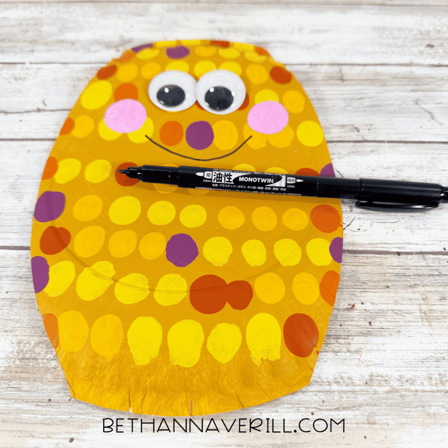 Dotted paper plate face with googly eyes, pink cheeks, and a curved smile being drawn with a black marker.