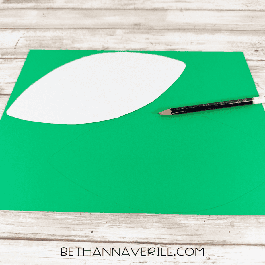 White leaf-shaped paper template resting on green paper with a pencil tracing another leaf shape.