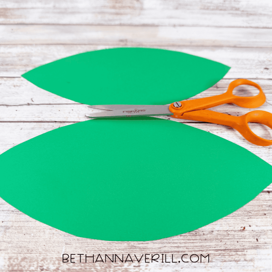 Two large green paper leaf shapes beside orange scissors on a white wood surface.
